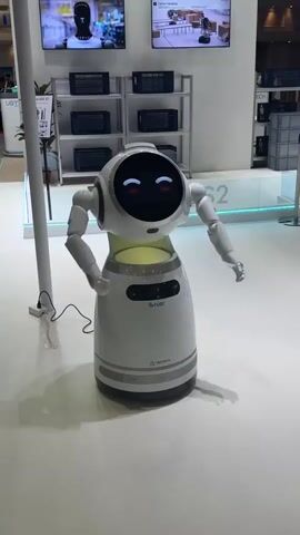 A white robot with a rounded body and a head with two circular digital eyes stands on a white floor. The robot's eyes are red and appear to be smiling. The robot has two articulated arms that are extended outward. Behind the robot, a shelf unit with several black appliances is visible, and above the shelf are two large screens displaying images. The environment appears to be an exhibition or trade show.