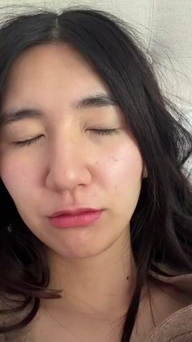 The video begins with a close-up of a woman's face as she opens her eyes. She has dark hair and light skin. The camera then quickly pans upwards to a window with white curtains. Green trees are visible outside the window. The shot then focuses back on the bed, covered with a white blanket.