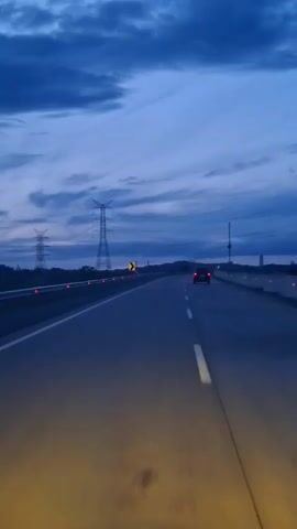 The video shows a view from inside a vehicle traveling on a highway at dusk. The sky is a deep blue with dark clouds. Tall power line towers are visible on the left side of the road. The highway has multiple lanes, and a single car is ahead in the right lane. Yellow headlights illuminate the road in the foreground. The road curves slightly to the right in the distance.