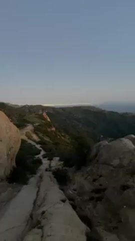 The video shows a landscape view from a rocky, mountainous area. In the distance, a city's lights are visible under a clear, pale sky. The foreground features large rocks and a narrow, winding path leading down through the green, hilly terrain. The light suggests it is dusk or dawn. There are no people visible in the scene.