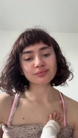 A close-up shot shows a young woman with short, curly brown hair and bangs, wearing a pink-strapped top with a leopard print. Her face is visible, and she is smiling slightly. A baby's hand, covered in a white sleeve or cloth, is resting on her chest. The background is a plain white wall. The camera angle is from below, looking up at the woman. The baby's hand moves slightly.