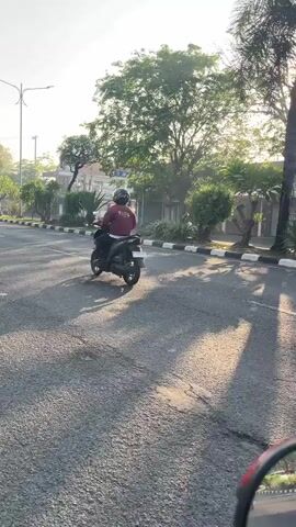 The video captures a street scene during daylight, likely morning or late afternoon due to the low angle of the sun casting long shadows. The camera appears to be mounted on a vehicle, possibly a motorcycle, as indicated by the reflection in a rearview mirror at the bottom right of the frame. The road surface is asphalt, with a distinct white and black dashed line marking the edge of the lane.

On the right side of the road, a dark-colored SUV is visible, parked or driving slowly. Further down the street, two motorcycles are in motion. The first motorcycle, a red one, is moving away from the camera, and the rider is wearing a helmet. The second motorcycle, to the right of the first, is also moving away, and its rider is also wearing a helmet.

The environment suggests a suburban or semi-urban setting. Trees with lush green foliage line the street, and the sunlight filters through their leaves, creating dappled patterns on the road. In the background, buildings with light-colored walls and tiled roofs are visible, along with some palm trees. The sky is clear and bright. The overall impression is one of peaceful, everyday activity on a sunny day.