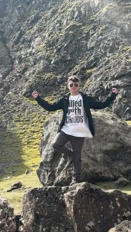 A young person stands atop a large rock, their arms outstretched, in a mountainous landscape. They wear a white graphic t-shirt, a dark jacket, and sunglasses.  The rock is surrounded by other rocks and some green mossy areas. Behind the person, a large rock formation towers, with a steep, rocky hillside covered with greenery. The camera zooms out to reveal the person standing among other rocks, with a larger formation in the background and a blue sky with puffy clouds overhead.