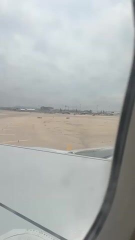 The video shows a view from an airplane window of an airport runway on a cloudy day. Several other airplanes are visible in the distance. The wing of the plane is visible in the lower left corner of the frame. The ground is concrete with yellow lines. There are several tall light posts and cranes in the background. The sky is overcast and gray. There's movement suggesting the plane is taxiing.