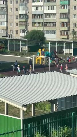 A group of young children, some wearing hats, are playing on a playground. A few adults, possibly supervisors or parents, are present. The playground is situated in front of a multi-story apartment building. The children are scattered across the playground, some running, others appearing to be in a line. One child is sitting on a bench or platform. The weather appears to be sunny.