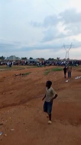 A large group of people, some on motorcycles, are gathered in an outdoor area with dirt ground and some vegetation. In the background, there are buildings and a tall electricity pylon. The sky is cloudy. A young boy in a patterned shirt and shorts is walking towards the camera in the foreground.