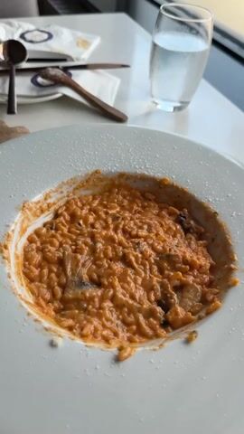 A close-up, top-down view of a white plate filled with risotto. The risotto is a vibrant orange color and appears creamy, with visible grains of rice. There are dark pieces mixed into the risotto, possibly mushrooms or other vegetables. Shredded white cheese is being sprinkled over the top of the risotto from the right side of the frame. In the background, a clear glass filled with water is visible, along with two wooden utensils and a white napkin. The scene appears to be set on a white table, likely in a restaurant.