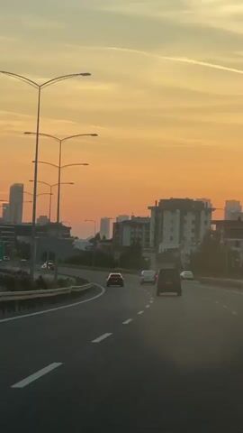 The video shows a view from a car driving on a highway towards a city skyline at sunset. The sky is a mix of orange, yellow, and light blue hues. Streetlights line the side of the highway. Several cars and a van are visible on the road. The city skyline features a mix of high-rise buildings and smaller structures. Some greenery is visible along the highway's edge. The camera is positioned inside the car, capturing the view through the windshield. There is also an audio component with the speech.