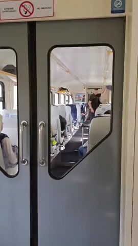 The video shows the interior of a train car from the perspective of looking through a glass panel in a door. The train car has rows of seats on either side of an aisle. Most seats are empty, but a few people are visible sitting down. A man in a dark shirt and shorts is walking down the aisle, carrying a duffel bag and a shopping bag. He is walking away from the camera. The train car appears to be in motion, as indicated by the slight blur of the windows and the context of being on a train.