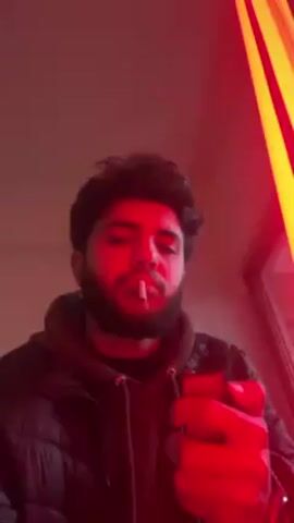 A young man with dark hair and a beard is seen from a low angle in a room lit with red light. He wears a dark hooded jacket and a watch on his left wrist. He lights a cigarette with a lighter, takes a drag, and exhales smoke toward the camera. A light fixture with red and yellow stripes is partially visible in the upper right corner.