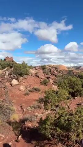 The video shows a landscape with red rock formations and scattered green shrubbery under a blue sky dotted with white, puffy clouds. A person wearing a white shirt, black shorts, a gray backpack, and neon green shoes walks away from the camera along a red dirt path. The landscape stretches into the distance, showing more rock formations and vegetation.