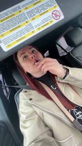 A woman with reddish-brown hair is sitting in the driver's seat of a car, applying lipstick. She is wearing a light-colored leather jacket, a black top with a white logo, and a tan seatbelt across her chest. She is holding the lipstick applicator near her lips, looking slightly upward. Above her, a sun visor is down, displaying warning labels in multiple languages. The car's interior is visible, including the black leather seats and the side windows. The camera is positioned at a low angle, looking up at the woman.