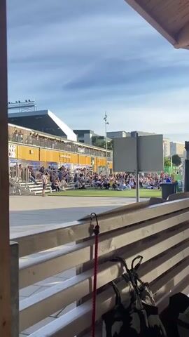 The video captures an outdoor gathering on a sunny day, likely in Uppsala, Sweden, given the context. A crowd of people are seated on tiered bleachers and spread out on a grassy area, suggesting a public event or performance. The architecture in the background, with its modern, modular design and yellow cladding, could indicate a venue or repurposed shipping containers. Signs visible on the building read "SUPPORTERS SHOP," implying a sports or fan-related event. The time of day appears to be late afternoon or early evening, as indicated by the warm, golden light and clear blue sky with scattered clouds. The overall atmosphere seems relaxed and communal, with people casually dressed and engaged in watching something happening out of frame. In the foreground, a wooden railing with some attached items like a red leash and bags suggests the viewer is observing the scene from a seated vantage point, perhaps a patio or deck area.