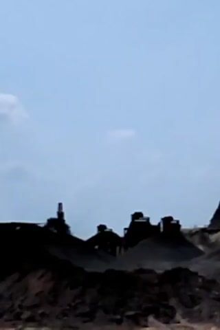 A distant view of an industrial site under a pale blue sky. The foreground consists of dark, uneven terrain, possibly piles of soil or excavated material. In the midground, silhouetted against the sky, is a cluster of industrial structures, including what appears to be a tall, slender tower or chimney with a red top, and several lower, blocky buildings. An excavator or similar piece of heavy machinery is visible to the left of the structures, its arm extended. The machinery and structures are dark, appearing as solid black shapes due to the lighting conditions and distance. The sky is mostly clear, with a few faint white clouds. The overall impression is of an active, industrial environment, likely a quarry or mine, viewed from a distance.