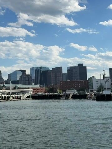 The video shows a view of the Boston skyline across the water.  The sky is blue with scattered white clouds.  Various buildings are visible in the skyline, including modern skyscrapers and older brick buildings. In the foreground, there is a pier with parking and several structures. The water is choppy with small waves. The camera appears to be moving slightly, likely from being on a boat.