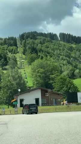 The video shows a ski lift ascending a lush green mountain covered in trees. In the foreground, there is a paved parking lot with a dark-colored van parked on the left side. Adjacent to the parking lot is a small building with white and wood siding. A sign with green text is visible on the left. There are people sitting on benches near the building. The sky is cloudy.