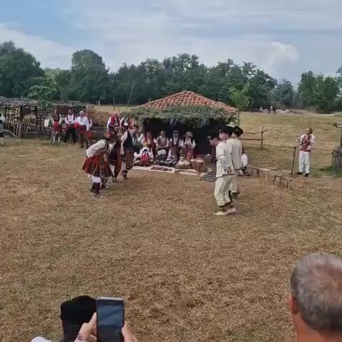 A wide outdoor shot of a cultural performance shows several people in traditional attire dancing in a circle on dry grass. They are performing in front of a small, open-sided structure with a tiled roof and leafy decorations, where other people in traditional clothing are seated, watching. To the right, another person in traditional attire stands near a wooden structure, possibly an old well. The background features scattered trees and a cloudy sky. In the foreground, the heads of two spectators are visible, one of whom is holding a smartphone, seemingly recording the event.