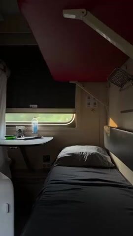 The video displays the interior of a train compartment, specifically a sleeping berth. A single bed is neatly made with dark grey sheets and a grey pillow, positioned in the lower right quadrant of the frame. To the left of the bed, a small fold-down table is attached to the wall. On the table, there is a clear glass, a partially full plastic water bottle, and a green object, possibly a pen. The window to the left of the table shows a blurred view of greenery outside, indicating the train is in motion. The compartment is dimly lit, with natural light entering through the window and artificial light from an overhead fixture, the red underside of which is visible at the top of the frame. The walls are a neutral, light color, and there are some electrical outlets and fixtures visible on the wall. The overall impression is one of a spartan but functional travel accommodation.