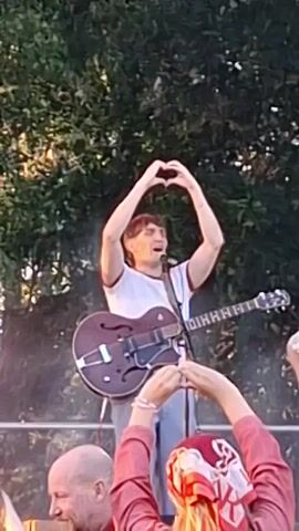 A man playing an acoustic guitar on stage is singing and making a heart shape with his hands above his head. A person in the foreground also makes a heart shape with their hands. The stage has a backdrop of trees and a speaker is visible. The lighting is dim with some smoke or haze effects.