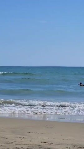The video opens with a shot of a sandy beach and the ocean on a clear day. The camera is positioned at ground level on the beach, looking out towards the water. Gentle waves are rolling in towards the shore. In the distance, on the right side of the frame, two people are visible swimming in the ocean. The water is a clear blue-green, and the sky is a bright, cloudless blue. The overall impression is a peaceful and serene beach scene.
