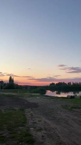 The video shows a wide shot of a rural landscape at dusk. The sky is a gradient of pale blue at the top to pink and orange near the horizon, with scattered clouds. In the middle ground, there is a body of water, likely a river or lake, reflecting the colors of the sky. Lush green vegetation lines the banks of the water. On the left side of the scene, there are some figures in the distance. One appears to be a person walking, and another might be an animal. Further left, there is what looks like the beginnings of a structure, possibly a building or fence. In the far distance, there are some trees and wind turbines. The foreground consists of a dirt path and some grass. The overall impression is of a peaceful, natural setting as day turns into night.