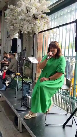 The video begins with a medium shot of a woman in a green dress singing into a microphone while seated on a stool. Behind her, a large, artificial white tree with blossoms is visible, along with a speaker and a sound system. The scene appears to be indoors, possibly a market or event space, with large windows and exposed brick walls in the background. A group of people are seated at tables in the background, some appearing to be eating.

The camera then pans to the right and focuses on a stall with a sign that reads "EL TORO" and features a red bull logo. Several people are standing in line at the stall. The video continues to pan across a lively indoor market environment, showing various food stalls, people browsing, and decorative flags hanging from the ceiling. The camera captures a sense of activity and community within the space, with individuals and groups interacting and moving through the market. The video ends with a wide shot of the bustling market, highlighting the vibrant atmosphere.