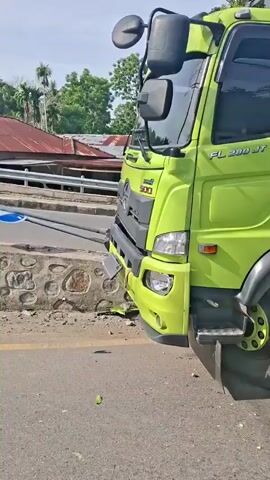 A bright green Hino truck has partially driven off a road and is resting against a stone wall. Debris is scattered on the road near the truck's front wheel. A motorcycle is visible passing the scene, with a person wearing a helmet. In the background, there are houses, trees, and a billboard. The sky is clear and blue.