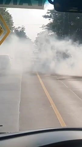 The video begins by showing a view from inside a vehicle looking forward. Thick white smoke billows across the road, obscuring several cars and the surrounding environment. The smoke appears to be coming from the tires of a vehicle on the right side of the frame, suggesting a burnout. The road is paved and has a yellow dividing line. Trees line the sides of the road. The perspective then shifts to a view from the side mirrors of the vehicle, showing more of the smoke. Finally, the video shows a view of an Indonesian flag flying in a rural or semi-rural area, with fields and some structures in the background. The image quality is poor and blurry throughout the video.