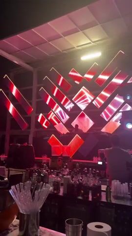 The video pans across a dimly lit bar area with bottles of alcohol and drink garnishes on display. In the background, a DJ is visible at a console in front of a large, dynamic LED screen featuring geometric patterns in shades of red, pink, and white. The ceiling above the stage has a grid-like structure with purple lighting. The overall atmosphere suggests a nightclub or event setting.