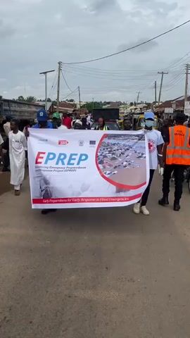 A group of people are marching down a paved street holding a large banner. The banner is white with red and blue accents and features the text "EPREP" and "Advancing Emergency Preparedness for Early Response Project (EPREP)". The banner also displays an aerial image of a flooded area with numerous houses submerged in brown water and tents set up on higher ground. The people holding the banner are wearing various clothing, including what appears to be a uniform with a red cross logo on the right side of the banner. A person on the far right wears an orange vest with "CIVIL DEFENCE" written on the back. In the background, other individuals are visible, along with buildings, vehicles, and utility poles. The sky is overcast.