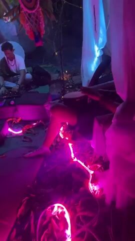 The video depicts an intimate, ambient music gathering set up outdoors. The scene is lit with a combination of pink and blue lights, creating a mystical atmosphere. A person is playing a small drum, while another individual sits cross-legged operating electronic music equipment. Around them, a small group of people are seated, enjoying the music. The area is decorated with fabric, dreamcatchers, and string lights. The ground is covered with mats and rugs, adding to the cozy vibe. Occasional glimpses show the natural outdoor environment, like trees and foliage, complementing the artistic setup.