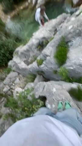 The video captures an outdoor scene in a rocky canyon or valley, likely Zupci, Montenegro. A person, dressed in a light-colored top and light bottoms, is seen bending down near the edge of a clear, light green/turquoise river or stream that flows through the landscape. The river is surrounded by large grey rock formations and lush green bushes and trees. In the background, steep, rocky mountains rise under a bright sky. Another person's legs and feet, wearing light blue or green footwear, are visible in the immediate foreground, suggesting the video is shot from a first-person perspective. The camera initially focuses on the person by the water and then quickly pans across the rocky terrain and water. The overall setting is natural and rugged, with no discernible text or specific time of day beyond daylight.