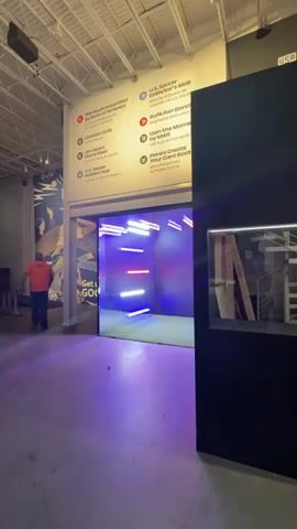 The video shows an indoor space, likely an exhibition or event venue, with a modern industrial aesthetic. The walls are white and high, with exposed ceiling beams and ductwork. The floor is concrete.

In the foreground, a person in an orange jacket walks away from the camera towards the left. To the right, a large, dark, rectangular structure dominates the view, with a horizontal illuminated window that reflects light.

Further into the space, a brightly lit entrance glows with neon-colored lights, predominantly pink and blue, arranged horizontally within a confined space. A sign on the wall above this entrance has text, some of which is visible: "GO!" and "GET".

To the right of the dark structure, the scene transitions to a display area with white walls. A black rolling tool cart with the brand "RIDGID" is visible, containing a green crate and other items. Behind it, there's a table and some cardboard boxes, suggesting ongoing setup or dismantling.

Further to the right, a red backdrop features the "BANK OF AMERICA" logo. The area is sectioned off with stanchions and velvet ropes. Another section has a blue wall with colorful graphics of a soccer ball and stars, and red and white striped walls. A small sign is visible on a velvet rope barrier. The overall impression is of a space being prepared for or currently hosting an event, possibly related to sports given the soccer ball imagery and "U.S. Soccer" text visible on a sign.
