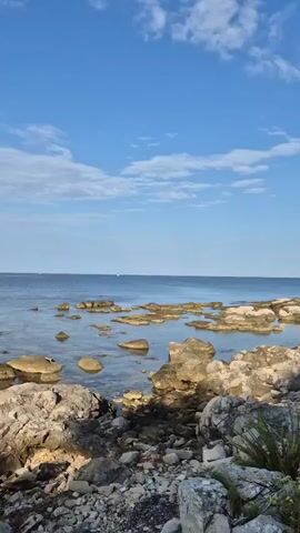 The video shows a rocky shoreline with clear blue water. The water is calm and shallow in some areas, revealing rocks beneath the surface. Larger rocks and pebbles are visible along the shore in the foreground. The sky is a vibrant blue with scattered white clouds. The scene suggests a peaceful, natural environment.