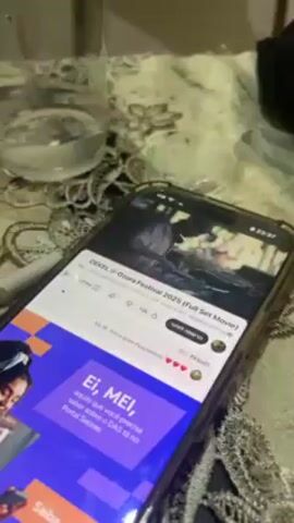 A close-up shot shows an iPhone displaying a YouTube video titled “DEKEL @ Ozora Festival 2023 (Full Set Movie).” The video itself shows a musician performing. An advertisement is displayed below the video with text in Portuguese. A glass of water is visible to the top left of the phone, and what appears to be a cigarette is to the left of the phone as well. The phone rests on a table with a textured, lace-like tablecloth. The lighting in the scene is somewhat dim, casting shadows on the objects.