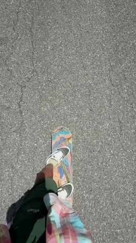 The video opens with an overhead shot of someone skateboarding on a paved road, wearing dark pants and a pink and white plaid shirt. The skateboard has a colorful, abstract design. The camera then shifts to a first-person perspective, looking down the road as the person continues to skateboard. A teal car is visible in the distance, driving away. The road is surrounded by lush green hills under a bright blue sky with white clouds. The shot returns to the overhead view of the skateboarder for a brief moment, showing their feet on the skateboard. The camera then returns to the first-person perspective, following the teal car as it drives further down the road, eventually disappearing from view. The landscape remains the same, with green hills and a clear sky.