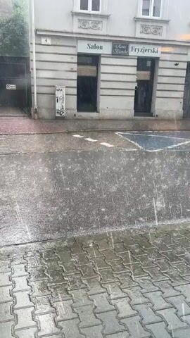 The video shows a street during a heavy downpour. Raindrops are visible splashing onto a paved sidewalk and a wet street. In the background, there is a building with several windows and signs, one of which reads "Salon Fryzjerski." A silver car is parked on the street. The scene appears to be in an urban environment.