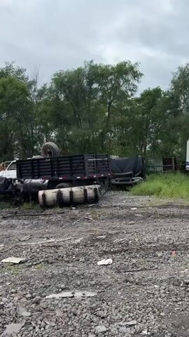 The video shows a junkyard filled with various types of vehicles and vehicle parts. In the foreground, there is a rocky and uneven ground with scattered debris. Several trucks are visible, including a large black flatbed truck with a tire on top, a white truck with a boom lift or bucket, and a white box truck. Other vehicles and parts appear to be piled up or dismantled. The background consists of trees and an overcast sky. The overall scene depicts a place where vehicles are stored for scrap or salvage.