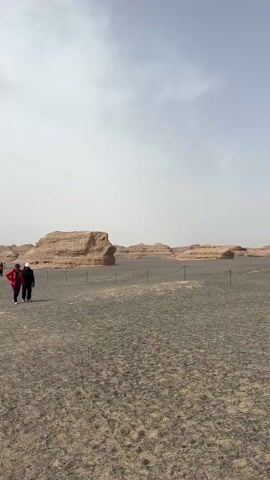 The video shows a wide, flat, gravelly landscape under a hazy, overcast sky. The scene is populated with several groups of people who are walking and taking pictures. In the distance, there are several large, natural rock formations, some with distinct horizontal layers, that resemble natural sculptures. A black rope line, supported by short posts, runs through the foreground, seemingly marking a boundary or a designated path for visitors. The overall atmosphere is dusty and muted in color, suggesting a dry, arid environment.
