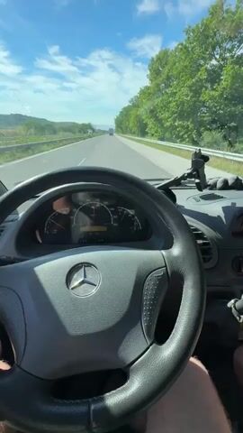 The video shows the interior of a vehicle, likely a Mercedes-Benz Sprinter van, as it drives on a highway. The camera is positioned behind the steering wheel, looking forward. The steering wheel, dashboard with gauges, and a portion of the passenger side are visible. The road ahead is clear, with trees lining the right side and a blue sky with clouds above. Other vehicles can be seen in the distance. The person driving is wearing shorts, and their hands are on the steering wheel. The video appears to be a first-person perspective recording of a road trip on a sunny day.