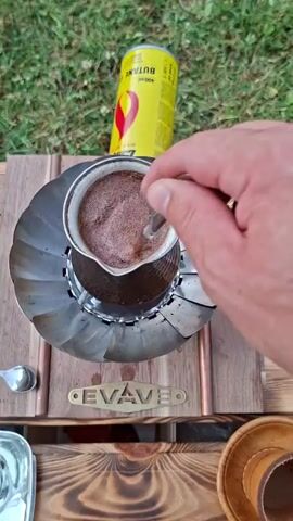 A person's hand is stirring coffee in a cezve with a spoon. A yellow butane gas canister is visible in the background. The cezve is on a metal stand, which is on a wooden surface. Another person's legs are visible in the background. The scene appears to be outdoors, with grass in the background.