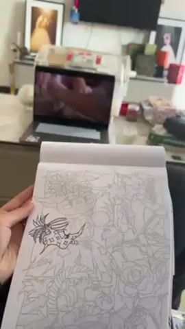 A person is shown holding a partially inked drawing in a sketchbook. The drawing appears to be a complex fantasy illustration with a character surrounded by flowers and foliage. In the background, a laptop displays a video where hands work with a brown, granular substance, possibly sand or soil, and a small tool. The laptop sits on a light-colored table. The background also shows framed photos, a TV screen, and various decorative items on the table. The camera angle focuses on the drawing and the laptop screen, providing a general overview of the scene and indicating that the person is likely drawing while watching the video.