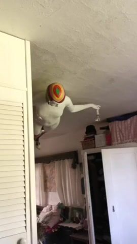 A person dressed in a full white bodysuit with metallic silver boots and a colorful target-design helmet is suspended upside down from a textured white ceiling. The individual holds a stemmed clear glass containing liquid, which also remains suspended without spilling. Below, a cluttered room is visible, featuring white louvered doors on the left, white curtains over windows in the background, a white wardrobe on the right, and various items including a bicycle and clothing scattered across the floor/furniture.