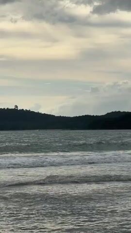 A serene beach scene unfolds under a cloudy sky. Gentle waves roll towards the shore, creating a soft, rhythmic sound. In the distance, lush green hills form a picturesque backdrop. A few individuals can be seen enjoying the water, with one person swimming and another on a surfboard. Further out, two colorful parasails drift through the air, adding a touch of vibrant activity to the tranquil setting. The overall atmosphere is one of peace and relaxation, inviting viewers to imagine themselves at this idyllic coastal escape.