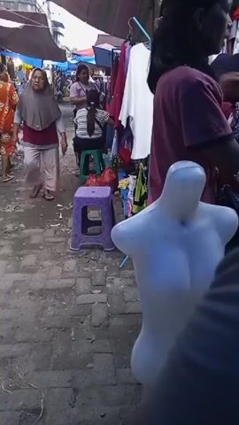 A busy outdoor market scene is captured. In the foreground, a white female mannequin torso is visible, positioned at an angle. The background reveals a bustling marketplace with several individuals. To the left, a woman wearing a grey hijab and a maroon top walks away from the camera, her body angled towards the left side of the frame. She is wearing light-colored pants and flip-flops. Further back, other shoppers and vendors are present. A woman in a colorful orange dress is walking away from the camera towards the left. Another woman is seated on a green stool, facing away from the camera, wearing a striped shirt and dark skirt. She appears to be interacting with another person behind her. A purple plastic stool is placed in the middle ground. Red plastic bags are visible near the green stool. Stalls with awnings are set up, displaying clothing and other goods. The ground is paved with stones and appears somewhat dirty. The overall environment suggests a lively, outdoor market setting.
