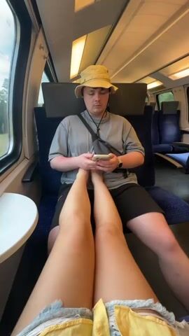 A person in a yellow bucket hat and grey t-shirt sits in a train seat looking at a smartphone. The person has their legs crossed and a woman’s legs are visible resting on their lap, almost between their hands holding the phone. The woman is wearing yellow denim shorts. The train car is mostly empty and it is daytime.