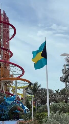 The video shows a sunny day at a waterpark.  The flag of the Bahamas waves from a white pole in the foreground. Behind it are several colorful waterslides, including a large red and orange one. Palm trees and other greenery are visible near the base of the slides.  A blue archway with "Thrill Waterpark" on it is in the bottom left of the frame. People walk near the entrance. Another tower with slides is visible on the right side. The sky is partly cloudy.
