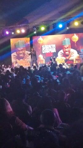 A live performance is taking place indoors in a large, crowded venue. An individual in a dark suit is on stage, holding a microphone and speaking. Two large screens behind him display the same image, featuring a smiling man wearing a hat, with the words "SARKIN DARIYA" above him. The lighting is a mix of blues, yellows, and purples. The audience sits at tables.