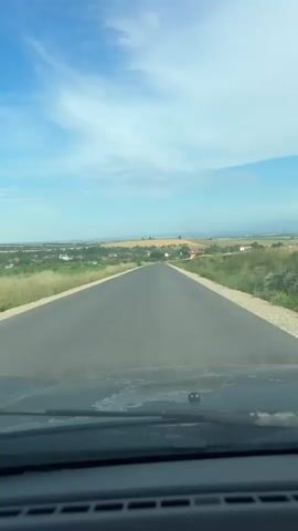 The video shows a POV shot from inside a car driving on an asphalt road. The road extends into the distance, leading towards a horizon with a village and rolling hills under a blue sky with white clouds. The foreground shows the car's dashboard and windshield. The scenery is rural, with green fields and some trees lining the road. The sky is bright, indicating daylight.