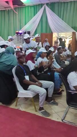 The video shows a gathering of people in a large room. Most of the people are seated in white chairs. Some of them are wearing white shirts and white caps, while others are wearing traditional attire including hijabs. A woman in a red patterned outfit wears a blue face mask. A woman in a black abaya and green head covering walks between rows of chairs. A red carpet runs through the center of the room. The room is decorated with green and white fabric draping in the background. Some people are dancing, and some are clapping and raising their hands in the air, possibly enjoying music that is playing in the background.